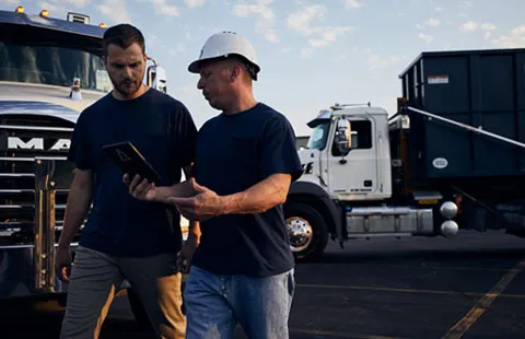 Two Men Talking While Walking Past Roll Off Dumpster Trucks